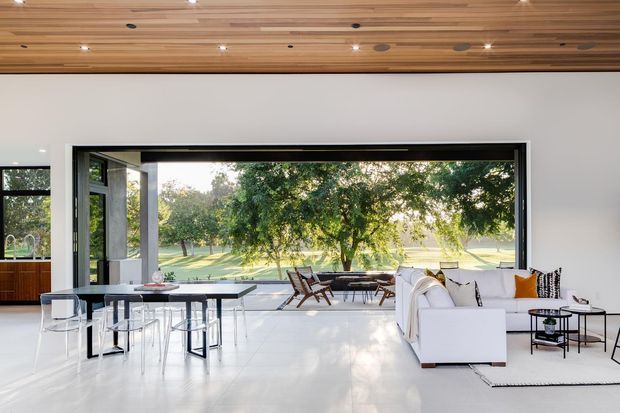 A bright, open-plan contemporary living and dining area with expansive black-framed glazing overlooking a leafy landscape. The room features a white sectional sofa, black dining table with transparent chairs, mid-century style lounge chairs, a low coffee table, and a striking slatted timber ceiling with recessed spotlights. White walls and glossy pale flooring create an airy, high-contrast interior.