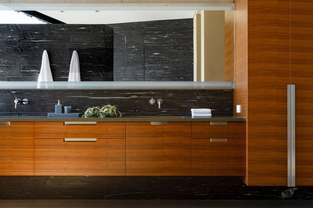 Contemporary bathroom vanity with extensive dark stone wall and splashback surfaces, warm timber veneer cabinetry, a long horizontal mirror, double wall-mounted faucets and integrated basin area. The room has a refined, high-contrast material palette with concealed storage and a large full-width mirrored wall.