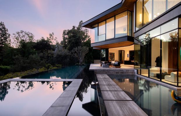 Exterior twilight view of a contemporary multi-level house set among mature trees, featuring extensive floor-to-ceiling glazing, projecting cantilevered upper volumes, dark horizontal cladding, timber soffits, and a series of reflective pools connected by broad stone or concrete stepping surfaces. A furnished outdoor terrace and warmly lit interiors are visible through the glazing.