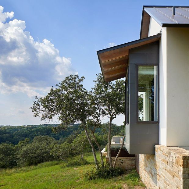Exterior architectural photograph of a contemporary multi-level home on a wooded, elevated site. The facade combines smooth white rendered walls, dark grey vertical cladding, a projecting sloped roof with exposed timber soffit, large vertical glazing, and a substantial light natural-stone masonry base. The house overlooks a heavily wooded landscape beneath a bright blue sky.