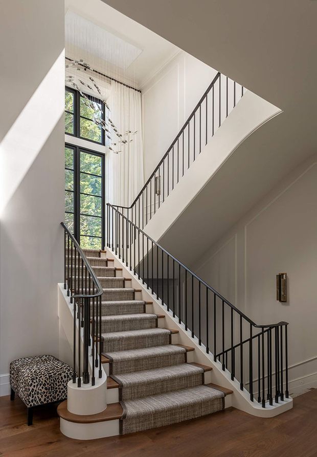 A bright, elegant residential stairway with a carpeted staircase, dark slender metal balustrades, white paneled walls, tall black-framed windows, and warm timber flooring. A small upholstered bench and sculptural pendant add decorative detail beneath the double-height stair hall.