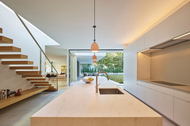 A bright contemporary open-plan kitchen viewed from the island, featuring a large pale stone island with integrated sink and brass-toned tap, extensive white flat-panel cabinetry, concealed lighting, pendant lights, and a sculptural floating timber staircase with glass balustrade. Floor-to-ceiling glazing opens toward a leafy outdoor area, while a skylight brings additional natural light into the space.
