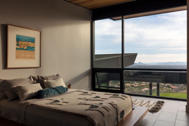 A contemporary bedroom with a low-profile bed dressed in neutral textured bedding and layered cushions. A framed artwork hangs on the warm grey wall, while expansive black-framed glazing provides sweeping views over a coastal or mountainous landscape. The room features a timber-lined ceiling, pale tiled flooring, and strong natural light.