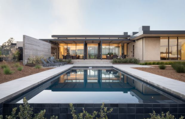 Wide exterior view of a low-slung modern residence arranged around a rectangular swimming pool. The home has extensive floor-to-ceiling glazing, flat rooflines, dark metal framing, pale rendered walls, a concrete or stone-paved pool surround, and a dark tiled pool edge. Warm interior lighting and furnished living spaces are visible through the glazing, while sparse landscaped planting and a rendered privacy wall frame the courtyard.