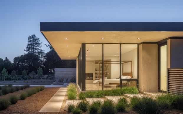 Twilight exterior view of a low-slung contemporary house with a broad flat roof and deep timber-toned soffit. Full-height glazed walls reveal a bedroom inside, while a paved path, ornamental grasses, timber screening, and a reflecting pool frame the modern facade.