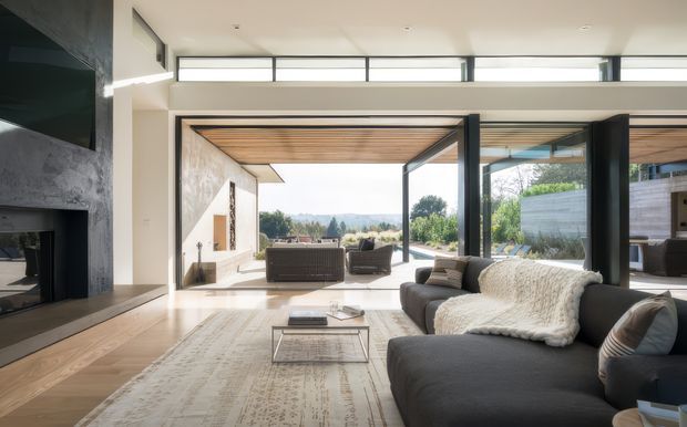 A bright, spacious contemporary living room opening onto an outdoor terrace through large retractable glazed doors. The room features a dark sectional sofa, pale patterned rug, low glass coffee table, minimalist fireplace wall with dark stone or concrete finish, extensive clerestory glazing, and a timber-lined ceiling continuing toward the outdoor area. The setting beyond the glazing includes landscaped grounds and distant hills.