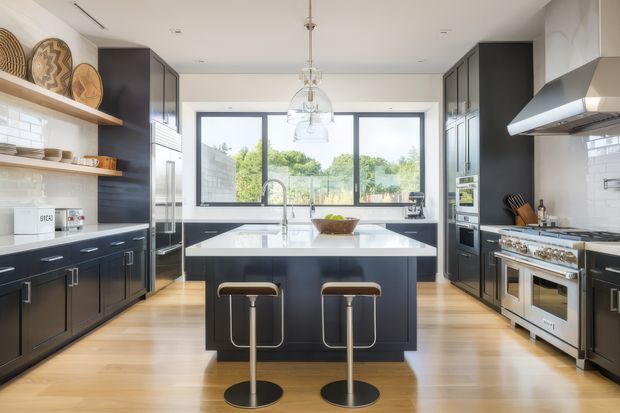 A spacious, contemporary kitchen viewed straight on, centred around a dark island with two stools. The room features dark built-in cabinetry, pale timber flooring, white benchtops, a white tiled backsplash, integrated appliances, a large professional-style range cooker, pendant lighting, and broad black-framed windows overlooking greenery.