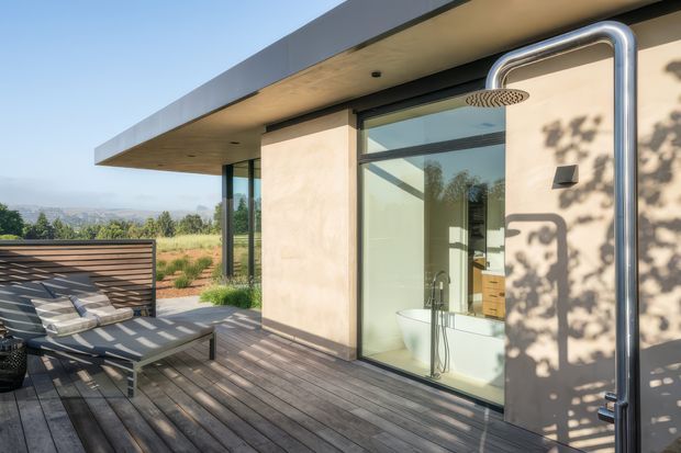 Exterior view of a contemporary home showing a timber deck and outdoor lounge beside a glazed bathroom. The bathroom has a freestanding bathtub, wall-mounted shower, and large sliding glass opening. The building features a flat roof, smooth light-toned walls, dark metal framing, and an outdoor stainless-steel shower. A landscaped property and distant landscape are visible beyond the deck.