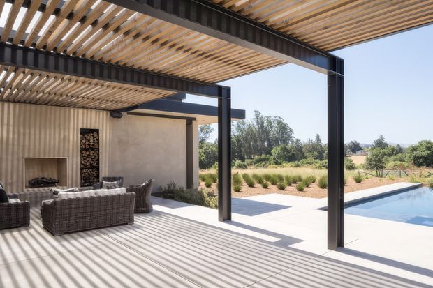 A modern covered outdoor living area with a timber slatted pergola roof, dark steel structural beams and columns, and a concrete or pale paved floor. Wicker lounge seating faces a built-in fireplace and stacked firewood niche. The space opens directly onto a rectangular swimming pool and landscaped garden, with strong striped shadows cast across the floor.