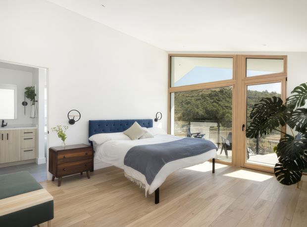 Bright contemporary bedroom with a low upholstered navy-blue bedhead, white bedding and a charcoal throw, positioned beside large timber-framed windows and a glazed door. Pale timber flooring, a mid-century-style timber bedside table, indoor plants and a partially visible adjoining bathroom create an airy, modern residential interior.
