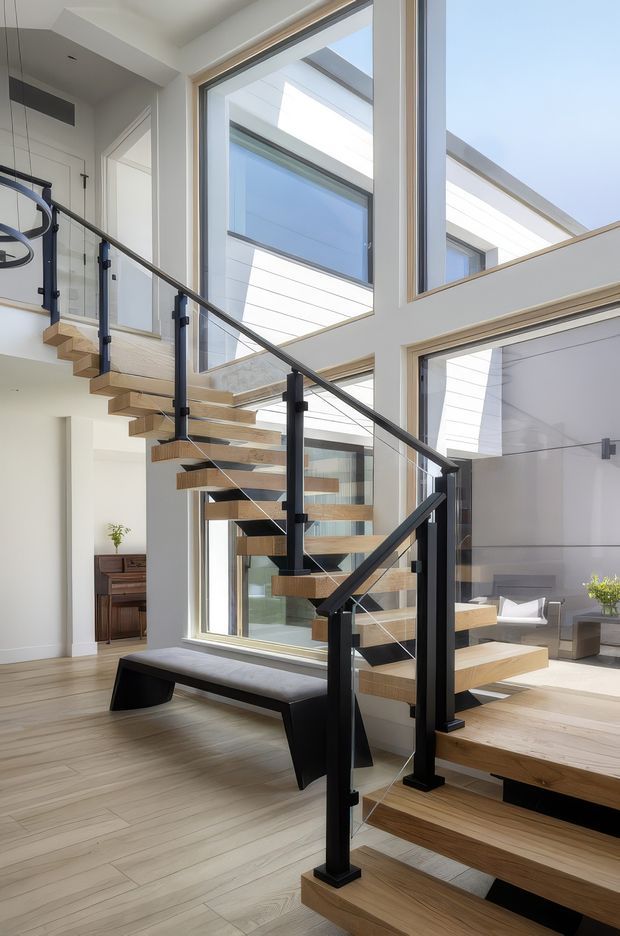 Bright contemporary interior centered on a floating open-riser timber staircase with black metal posts, handrail, and clear glass balustrades. The stair hall has light timber flooring, white walls, extensive floor-to-ceiling glazing, and a strong indoor-outdoor visual connection to a modern courtyard or adjoining space. A dark built-in bench sits beneath the stair, with a small timber console visible in the background.
