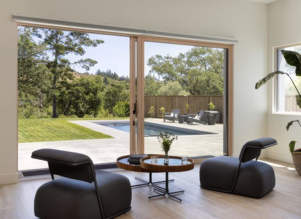 Bright contemporary interior seating area with two sculptural dark lounge chairs and a round coffee table, positioned in front of large timber-framed sliding glass doors. The doors open to a broad paved terrace, swimming pool, lawn, and landscaped garden beyond. A large indoor plant and a small side window add natural detail, while pale walls and light timber flooring create an airy, refined setting.