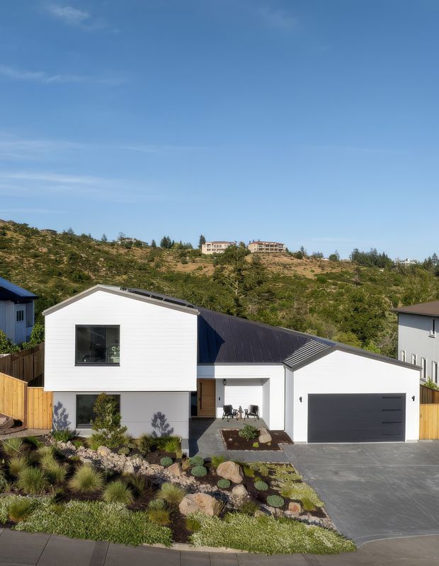 Street-facing exterior photograph of a contemporary two-storey white-clad house with a prominent asymmetrical gable roof, attached single-storey garage, dark metal roofing, timber privacy fencing, and a landscaped front garden with rocks and drought-tolerant planting. The home sits on a sloping suburban hillside with other houses visible in the background.