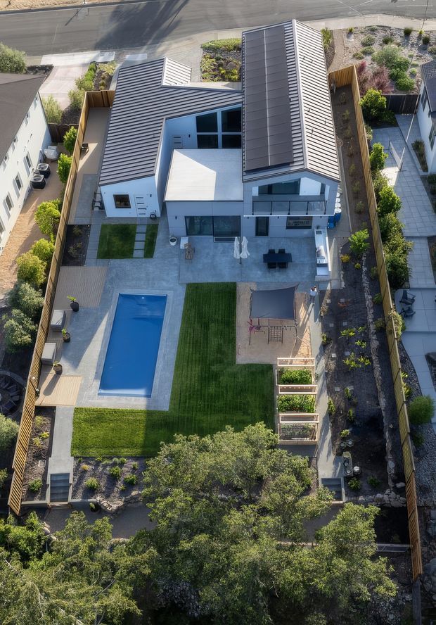 Aerial view of a contemporary multi-level residential property with a distinctive pitched metal roof, light-coloured rendered walls, and a landscaped backyard. The outdoor area includes a rectangular swimming pool, lawn, paved entertaining terrace, raised garden beds, children’s play equipment, timber fencing, and planted garden beds. The home is situated on a narrow suburban site alongside a paved street.