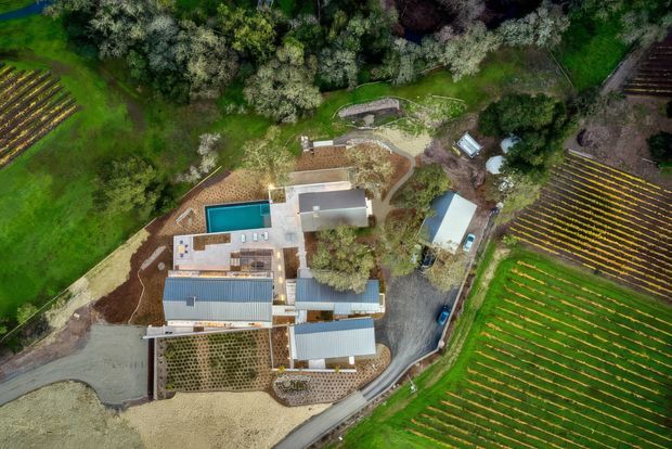 Aerial top-down view of a rural property surrounded by cultivated agricultural fields and dense trees. The complex includes several low-rise buildings with light and dark metal roofs, paved paths and courtyards, landscaped areas, and a rectangular turquoise swimming pool. The site appears integrated into a working vineyard or farm landscape.