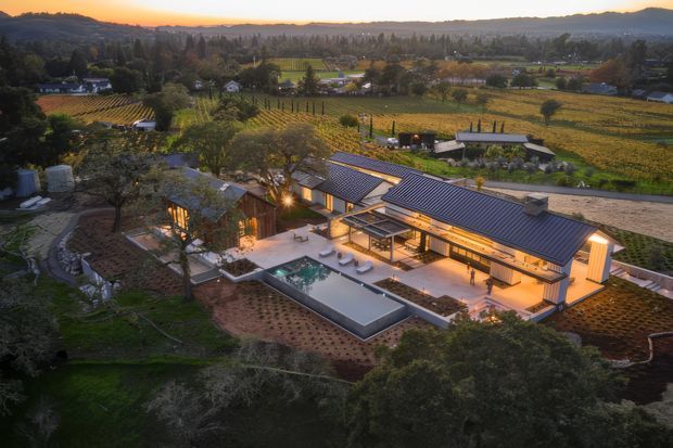 Aerial dusk view of a low-rise contemporary residence set within a cultivated rural vineyard or orchard landscape. The home has elongated pitched metal roofs, warm illuminated interiors, extensive glazing, landscaped courtyards, and a rectangular swimming pool with surrounding outdoor terraces. Rolling hills and agricultural fields form the background.