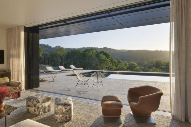 A refined living room opens through fully retracted floor-to-ceiling glazed doors onto a broad paved terrace and infinity-edge pool, with lounge chairs overlooking a wooded, rolling landscape. The interior features sculptural upholstered chairs, patterned ottomans, curtains, a textured area rug, and a dark contemporary ceiling and door frame system. Warm late-day light creates a sophisticated indoor-outdoor atmosphere.