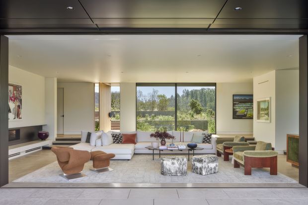 A bright, spacious contemporary living room viewed from a wide glazed opening. The room features a white sectional sofa, sculptural brown lounge chairs, a large pale rug, low round coffee tables, artwork, and built-in wall cabinetry. Floor-to-ceiling glazing opens the room to a leafy outdoor outlook, while a dark, paneled ceiling and dark framing create contrast with the light interior.