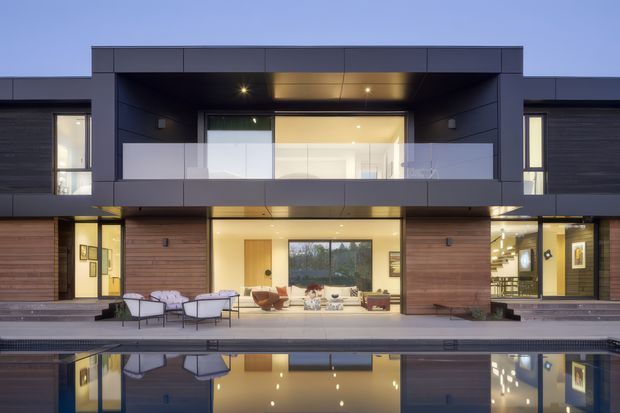 A dusk exterior view of a large contemporary multi-storey residence with a dark, rectilinear facade, warm timber wall cladding, extensive glazing, and a recessed upper-level balcony with a translucent glass balustrade. The ground floor opens onto a paved terrace furnished with outdoor seating and leads directly to a long reflecting swimming pool. Warm interior lighting and the pool reflection create a polished, luxurious atmosphere.