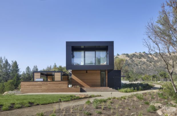 Exterior view of a contemporary two-storey residence set on a landscaped, sloping site. The house has a strongly geometric dark-clad upper volume with large glazing, a warm timber-clad entry and lower-level pavilion, and broad timber decking or terrace areas. Dry hills, mature trees, and open planting surround the property under a clear sky.