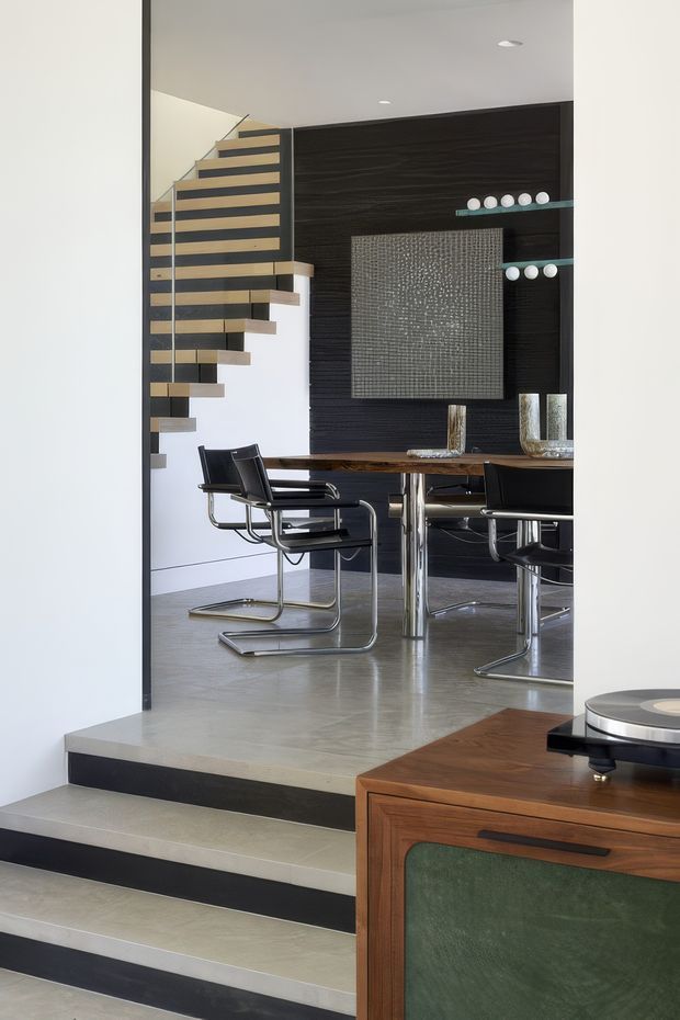 A refined modern interior viewed from a stepped entry area, looking into a dining room. The dining space features a dark timber-look feature wall, a large textured artwork or panel, glass display shelves, a polished rectangular dining table, and black cantilevered dining chairs with chrome frames. A floating open-riser staircase with timber treads and dark horizontal balustrade elements occupies the left side. The foreground includes a mid-century-style timber console with a green inset panel and a turntable. Neutral white walls, grey flooring, and strong black detailing create a sophisticated, high-contrast composition.