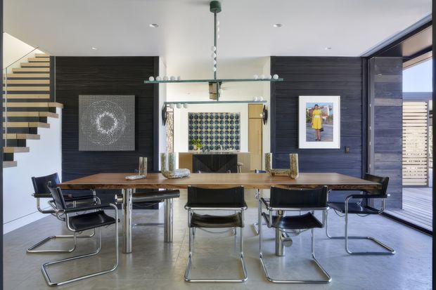 Contemporary dining area with a long timber dining table and black cantilever chairs, set against dark horizontal wall panelling. A floating staircase is visible at left, while a glass display shelf and framed artwork form the backdrop. Large glazing and an external louvre screen bring in daylight from the right.