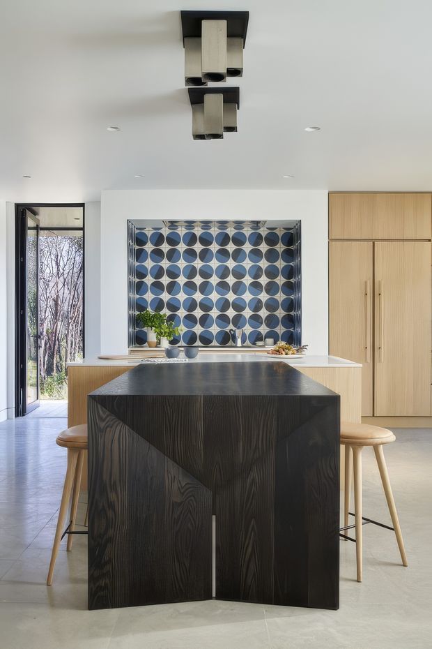 A bright, contemporary kitchen viewed from the main living area, featuring a dramatic black timber-grain island with sculptural angled panels and integrated seating. Behind it is a pale timber wall of tall cabinetry, a light benchtop, and a bold blue-and-white geometric tiled splashback with circular motifs. A glazed external door, pale floor, recessed ceiling lights, and black-and-metal pendant fixtures complete the space.