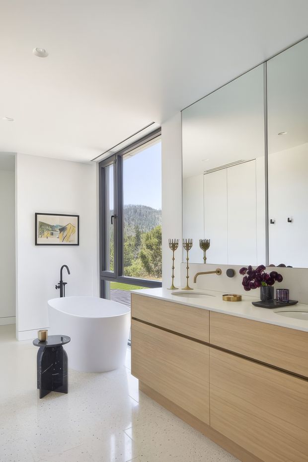 A bright, contemporary bathroom with a freestanding white bathtub beside a large window overlooking greenery and hills. A long pale timber vanity with integrated white countertop and basin runs along the right wall beneath a full-width mirrored cabinet. Matte black tapware, a tall black floor-mounted bath filler, brass decorative candleholders, and restrained accessories add contrast. The room has white walls, recessed ceiling lights, and a polished light terrazzo-style floor.