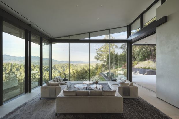 A sophisticated contemporary living room with floor-to-ceiling black-framed glazing on three sides, offering expansive views over a wooded, rolling landscape. The room features a large neutral sectional sofa, matching armchairs, layered round coffee tables, a dark textured area rug, recessed ceiling lights, and a raised opening toward an adjacent outdoor terrace or balcony. The space is spacious, bright, and strongly connected to the landscape.