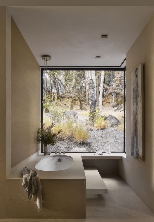 A serene minimalist bathroom with a sculptural integrated vanity and white vessel basin, pale beige wall finishes, a frameless black-edged picture window overlooking a wooded natural landscape, and a built-in low ledge or bench beneath the window. Simple pendant lighting and recessed ceiling fixtures create a calm, warm atmosphere.