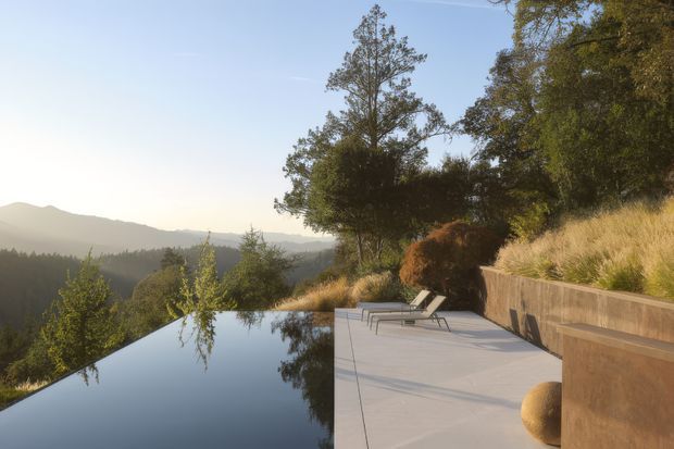 A serene elevated outdoor terrace beside a dark reflective infinity pool, overlooking wooded hills and distant mountains. The terrace has pale concrete paving, minimalist sun loungers, a sculptural rounded stone, and a built-in rust-toned retaining wall integrated into a dry grassland landscape. Warm late-afternoon light creates a calm, natural atmosphere.