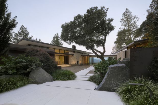 A low-slung modern residence viewed from a landscaped entry path. The composition shows flat-roofed wings, extensive warm-toned glazing, pale stone or masonry walls, dark metal elements, and large natural boulders integrated into a carefully planted garden. A central courtyard-like approach leads toward the house, with a distant blue view visible through the glazing.