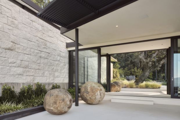 A contemporary covered entry or transitional outdoor space with broad open glazing and dark metal framing. A pale grey horizontally textured stone wall lines the left side, while large spherical stone ornaments sit on a smooth light floor beside planted borders. The space opens toward a landscaped garden and includes a dark slatted overhead canopy, creating a refined indoor-outdoor connection.