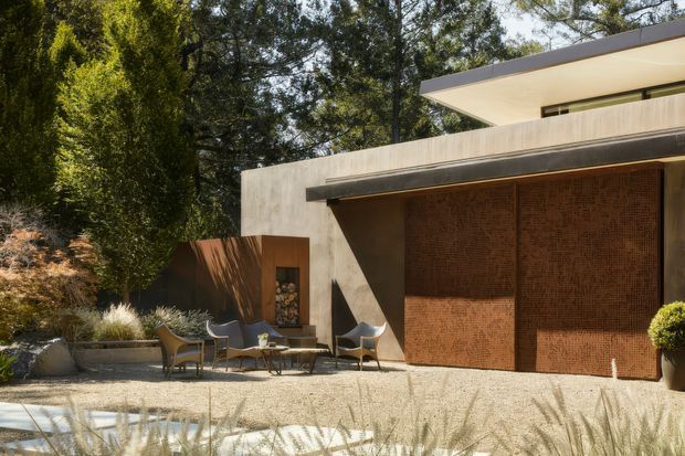 A refined modern residential exterior set within a landscaped garden. The low-slung structure features broad flat roof planes, smooth pale concrete or rendered walls, dark trim, and a large warm rust-brown textured feature wall or door. A gravel courtyard with outdoor lounge furniture and ornamental grasses sits in front of the building, surrounded by mature trees and sculpted planting.