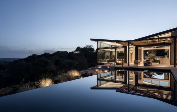 A dramatic dusk exterior view of a contemporary, multi-level home set above a dark landscaped hillside. Extensive floor-to-ceiling glazing opens to a covered outdoor living area with timber slatted screening and warm interior lighting. A highly reflective infinity-edge pool dominates the foreground, mirroring the house and sky.