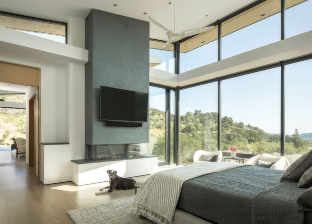 Bright contemporary bedroom with a high ceiling and expansive floor-to-ceiling corner glazing overlooking a wooded landscape. The room features a large bed, minimalist built-in fireplace and television wall, pale timber flooring, and a connected upper-level void or mezzanine visible above the glazing. A dog is present on the floor.