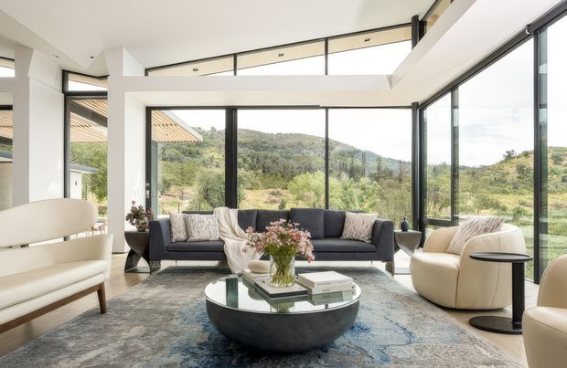 Bright contemporary living room with expansive black-framed floor-to-ceiling glazing overlooking a hilly, landscaped setting. The room features a dark sectional sofa, curved cream lounge seating, a sculptural round coffee table, a large patterned rug, and a high white ceiling with clerestory windows and recessed lighting.