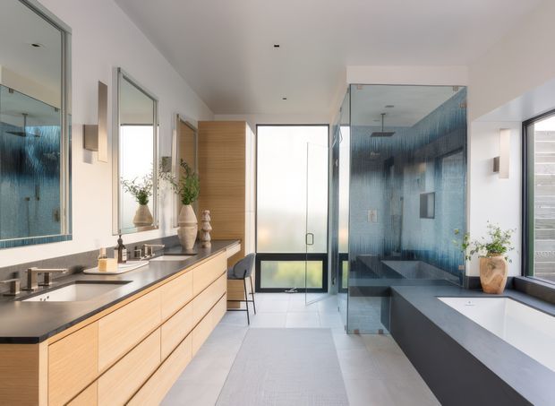 Bright contemporary bathroom with a long floating timber vanity, dark stone benchtop, large wall mirror, freestanding bathtub, and a fully glazed shower enclosure lined with blue textured tile. A frosted-glass door and clerestory-style glazing bring natural light into the room. The palette combines pale timber, white surfaces, charcoal grey, and blue tile accents.
