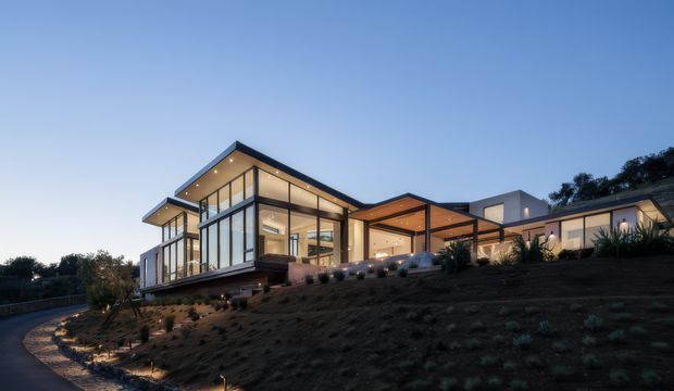 A contemporary multi-wing residence on a sloping landscaped site, photographed at dusk. The house features extensive floor-to-ceiling glazing, strong geometric volumes, projecting flat roof planes, covered outdoor terraces, warm timber soffits, and dark structural framing. Low landscape lighting illuminates the pathway and planting around the home.