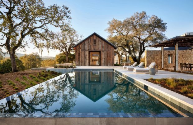Exterior view of a landscaped rural property centred on a long reflecting swimming pool, with a timber-clad gabled barn-like pavilion at the far end and a stone-clad open-sided structure to the right. Mature trees and rolling terrain surround the pool terrace.