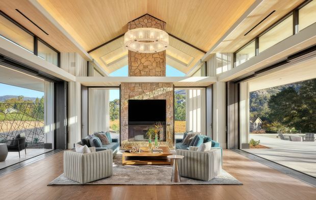 A spacious, bright contemporary living room with a dramatic double-height stone fireplace and chimney breast as the central feature. The room has extensive floor-to-ceiling glazing, exposed views to the surrounding landscape, a warm timber-lined vaulted ceiling, timber flooring, two sculptural upholstered sofas, lounge chairs, and a central coffee table. A large circular pendant light hangs in front of the fireplace, while an adjacent open terrace is visible through the glazing.