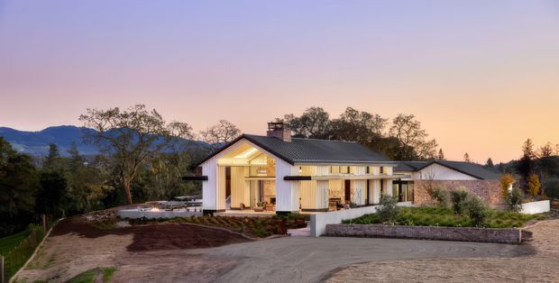 Exterior dusk view of a low-slung contemporary house set in a landscaped, elevated site. The home has a strong gabled central volume with large glazed openings, pale rendered walls, dark metal roofing, and long horizontal wings extending to either side. A broad driveway and terraced garden surround the building, with wooded hills visible in the background.