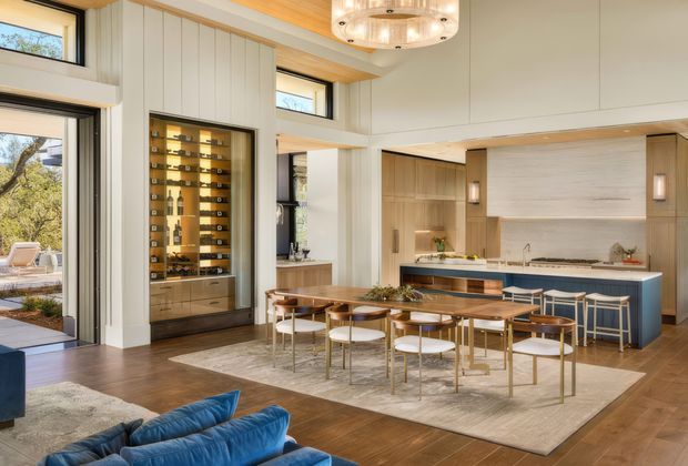 Open-plan kitchen and dining area with a large island, blue cabinetry, integrated appliances, a timber dining table, and multiple upholstered dining chairs. The room features warm timber flooring, light vertical wall panelling, extensive glazing, a built-in wine display cabinet, and a statement circular chandelier. A blue sofa is partially visible in the foreground, connecting the space to a living area.