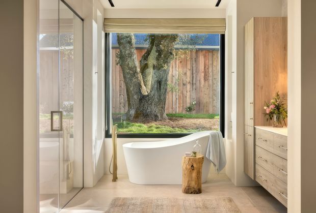 Bright contemporary bathroom featuring a freestanding white bathtub positioned beside a large glazed opening overlooking a private garden. The room has warm pale timber cabinetry, a timber feature wall, light neutral flooring, a frameless glass shower enclosure, recessed ceiling lighting, and natural wood accents.