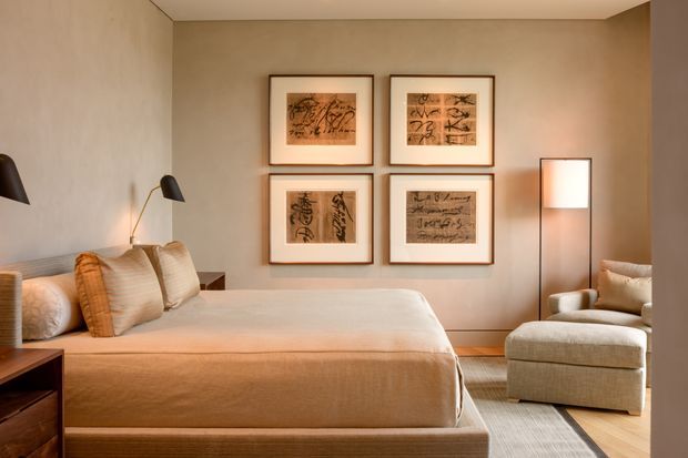 Warm, refined bedroom interior with a large upholstered bed, layered neutral bedding, bedside lighting and timber nightstands. A grid of four framed artworks decorates the beige feature wall, while a pale upholstered lounge chair and ottoman sit beside a slim floor lamp. The room has timber flooring, soft area rugs and a restrained neutral palette.