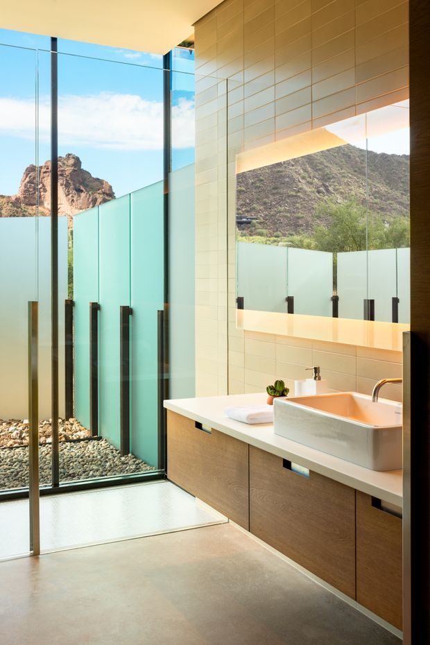 Bright contemporary bathroom with a walk-in glass shower, floating wood-look vanity, white vessel sink, large illuminated mirror, and pale tiled wall. Full-height glazing and frosted privacy panels open the space to a rugged mountainous landscape and blue sky. The room has a clean, warm-modern material palette with strong indoor-outdoor connection.
