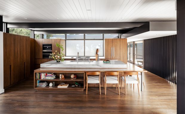 A spacious open-plan kitchen and dining area with a large white stone island, integrated timber shelving, dark timber cabinetry, stainless-steel appliances, and four timber bar stools. Full-height glazing and a glazed side wall bring in abundant natural light, while a dark exposed structural beam and vertical black slatted screen add strong contrast. The room has warm timber flooring and a white linear-panel ceiling.