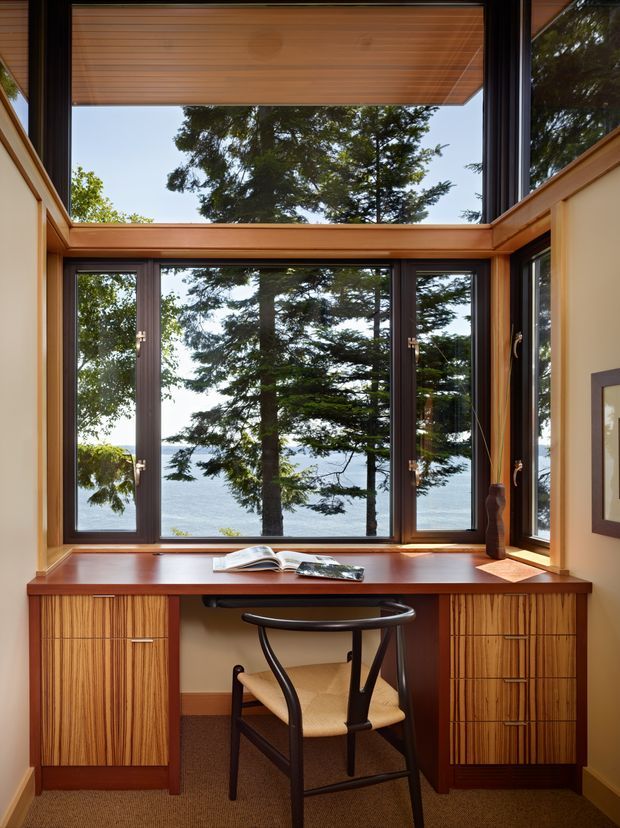 A compact built-in study nook positioned in front of expansive timber-framed windows overlooking trees and water. The space features a warm timber desk surface and matching vertical-grain built-in cabinetry, dark window frames, a simple black-and-cane chair, and a timber-lined overhead window reveal. Natural light and the view dominate the setting.