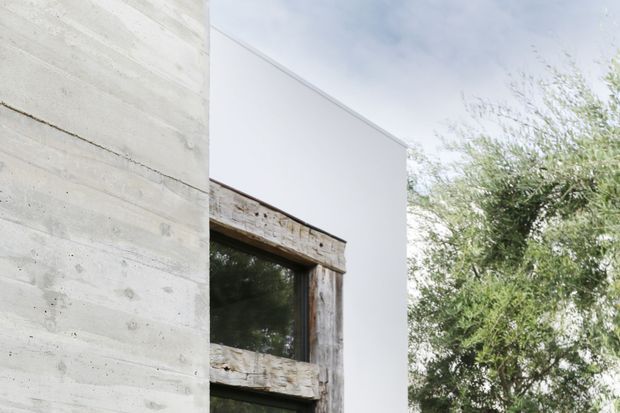 Minimalist exterior architectural detail showing a pale board-formed concrete wall beside a smooth white rendered volume and a large window framed in weathered reclaimed timber. Mature greenery is visible to the right, and the composition emphasizes contrasting raw and refined facade materials.