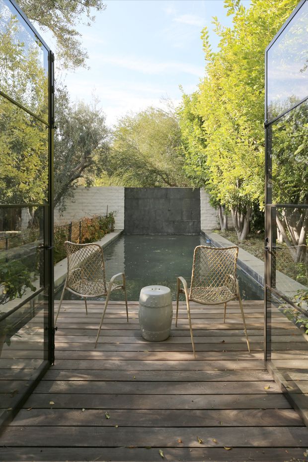 A tranquil outdoor pool courtyard viewed through fully open black-framed glass doors. A dark timber deck leads to a narrow rectangular pool enclosed by pale and dark masonry walls, with two woven outdoor lounge chairs and a small round ceramic side table. Dense mature planting surrounds the space, creating a private garden setting with soft natural light.